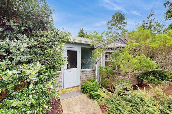 A small, single-story house with a front door, a visible window, and a pathway leading to the entrance.