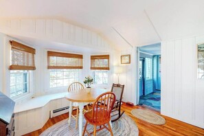 A dining area with a round table, wooden chairs, and a bench. There are three windows with bamboo shades, a lamp, and a view of a blue room through an open door. - Charming island cottage near town & beaches (Edgartown)