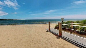 Une plage de sable avec une promenade en bois menant à l’océan, un panneau indiquant l’accès public à la plage et un ciel bleu dégagé.