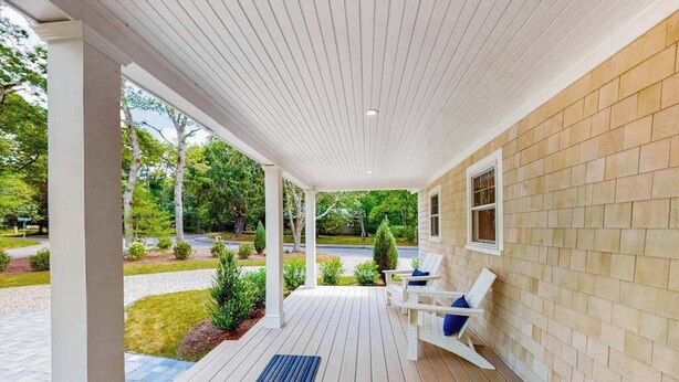 Une véranda couverte avec une terrasse en bois, des colonnes blanches et une vue sur un jardin aménagé.