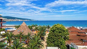 A beachside view with a thatched-roof structure, palm trees, and a clear blue sky. - Largo Beach Hotel (Sunny Beach)