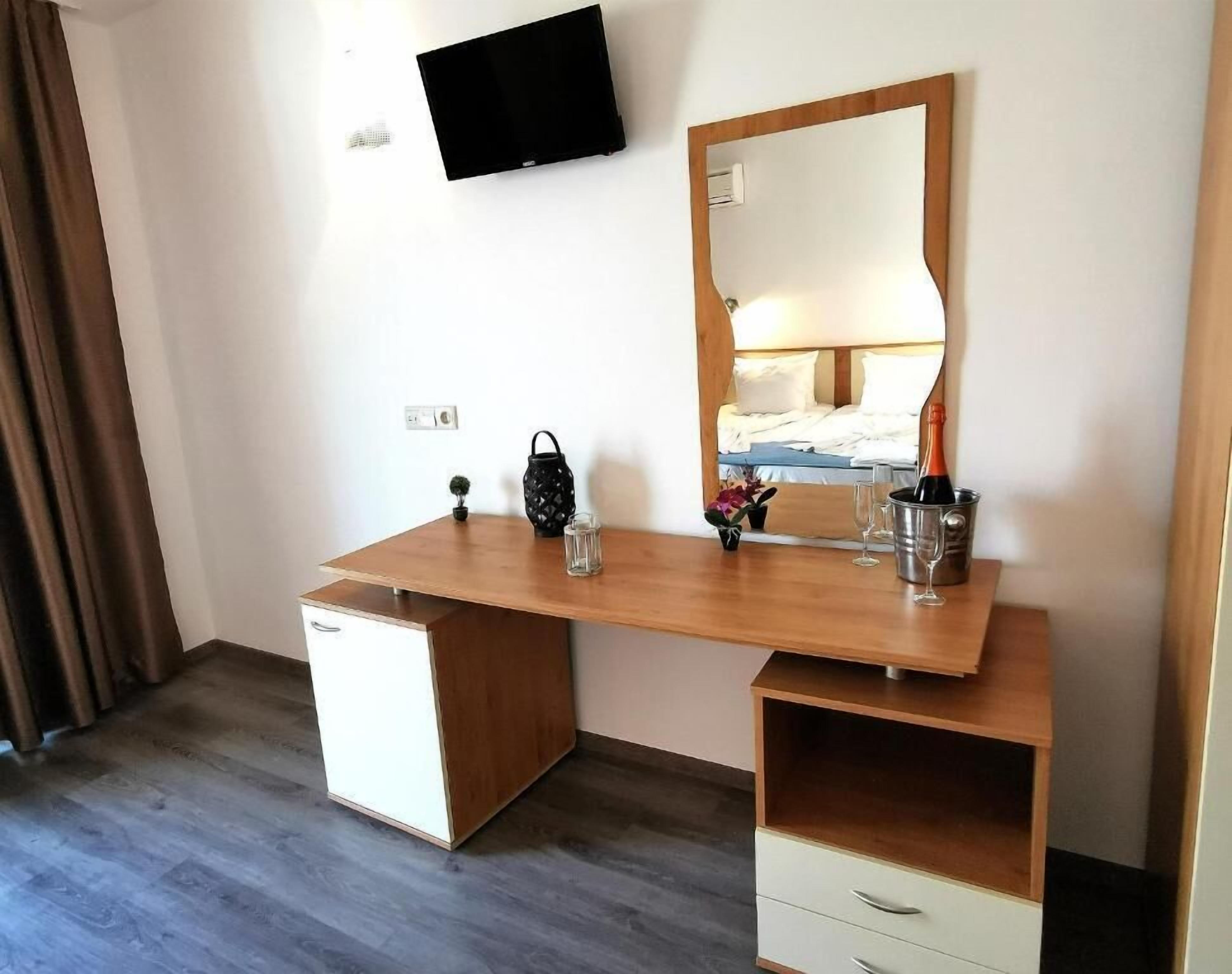 A hotel room with a wooden desk, a mirror, a television mounted on the wall, a bed with white bedding, and a small potted plant.