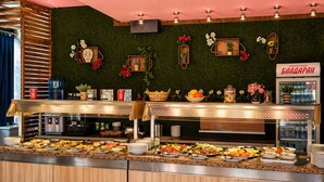 A buffet with various food items, a green wall with decorations, and a sign in a foreign language. - Largo Beach Hotel (Sunny Beach)