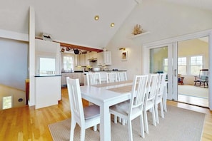 A modern kitchen with white cabinets, a dining area with a white table and chairs, and a large sliding glass door leading to an outdoor area. - Cape Beach House with spectacular views and beach access, central AC (Eastham)