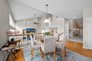 A spacious living room with a round glass table, white chairs, a ceiling fan, and a staircase leading to another level.