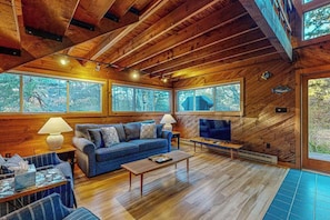 Interior de una cabaña de madera con sofá, televisión y ventanas amplias.