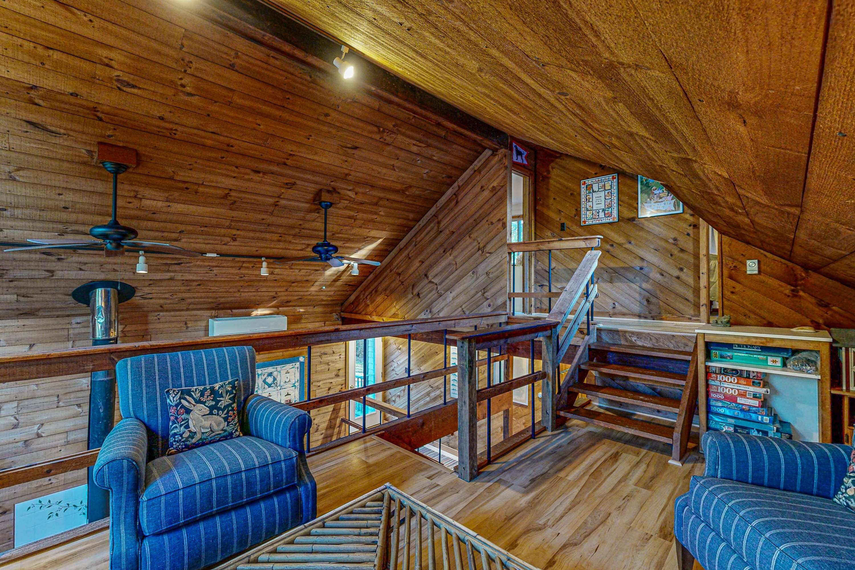 Un intérieur en bois avec un escalier, deux fauteuils bleus rembourrés, des ventilateurs de plafond et une bibliothèque.