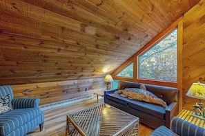 A cabin interior with a wooden ceiling, a sofa with a leopard print pillow, a striped armchair, and a window with a view of trees.
