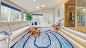 A spacious living room with a large sectional sofa, a coffee table, and a patterned rug. There are large windows, a ceiling fan, and a wooden staircase. - Expansive Getaway w/ Fenced Yard, Outdoor Shower & Private Neighborhood Beach (Hyannis)