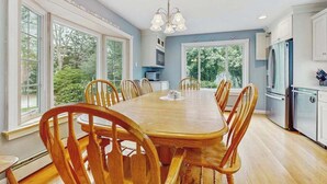 A dining room with a wooden table and chairs, large windows, and a kitchen area with appliances. - Expansive Getaway w/ Fenced Yard, Outdoor Shower & Private Neighborhood Beach (Hyannis)