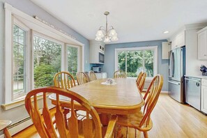 A dining room with a wooden table and chairs, large windows, and a kitchen area with appliances. - Expansive Getaway w/ Fenced Yard, Outdoor Shower & Private Neighborhood Beach (Hyannis)