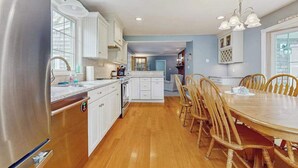 A modern kitchen with white cabinets, a wooden dining table, and a chandelier. - Expansive Getaway w/ Fenced Yard, Outdoor Shower & Private Neighborhood Beach (Hyannis)