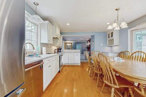 A modern kitchen with white cabinets, a wooden dining table, and a chandelier. - Expansive Getaway w/ Fenced Yard, Outdoor Shower & Private Neighborhood Beach (Hyannis)