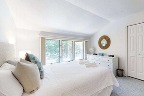 Ein Schlafzimmer mit einem großen Bett, einem Schminktisch, einem Spiegel und einem Fenster mit Blick auf Bäume.