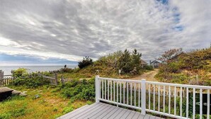A coastal view with a wooden deck, a white fence, and a path leading to a building. - Newly built, bayview house steps from the beach with spacious deck, W/D & grill (North Eastham)