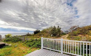 A coastal view with a wooden deck, a white fence, and a path leading to a building. - Newly built, bayview house steps from the beach with spacious deck, W/D & grill (North Eastham)