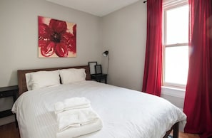 Un lit bien fait, avec du linge de lit blanc et une serviette pliée, un tableau floral rouge accroché au mur et une fenêtre ornée de rideaux rouges.