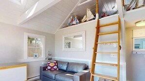 A room with a grey sofa, a wooden ladder, and a window. - Quiet cottage on the beach with deck, grill, AC, & water views (Truro)