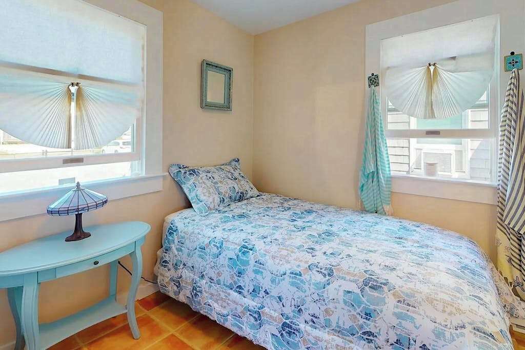 Quarto com cama, mesa e duas janelas com cortinas.