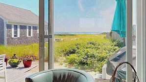 A view through a glass door to a deck with a chair, potted plants, and a grill. - Quiet cottage on the beach with deck, grill, AC, & water views (Truro)