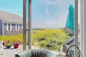 A view through a glass door to a deck with a chair, potted plants, and a grill. - Quiet cottage on the beach with deck, grill, AC, & water views (Truro)