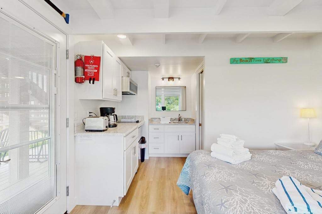 A hotel room with a kitchenette, a bed with a patterned bedspread, a bathroom with a mirror, and a sign on the wall.