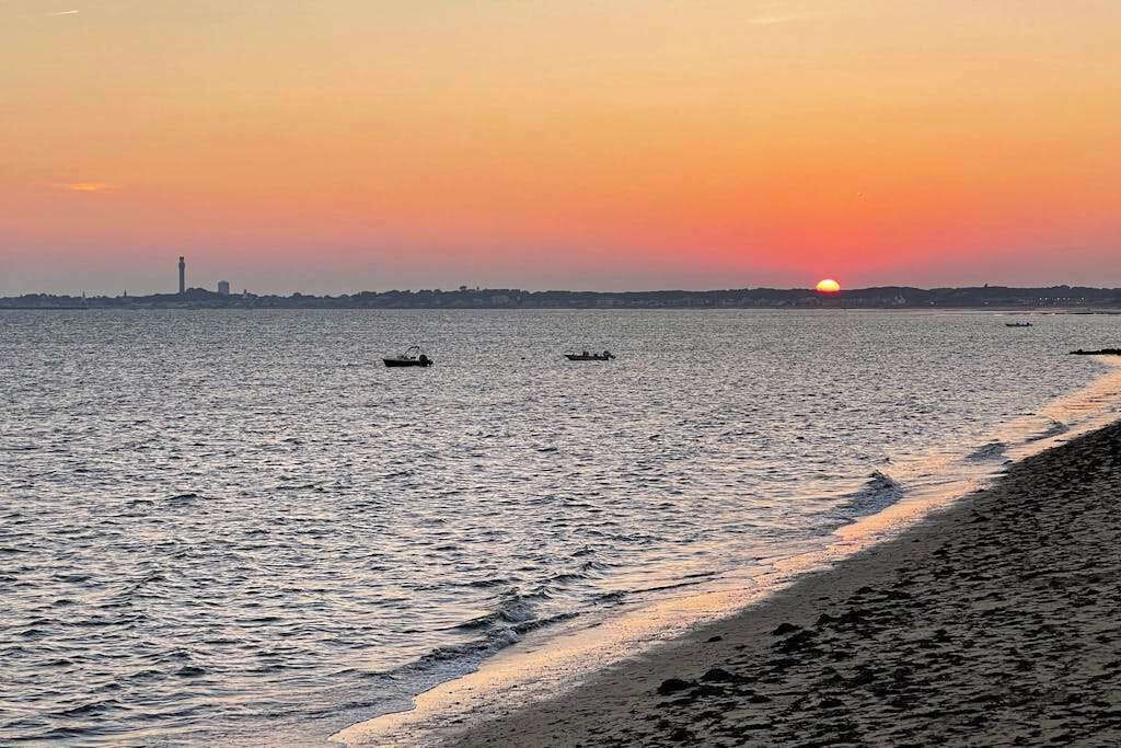 Solnedgang over et roligt hav med både, et fjerntliggende fyrtårn og en strand.