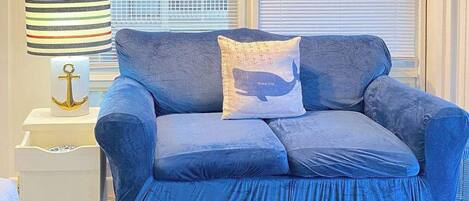 Un canapé bleu orné d’un coussin en forme de baleine, une lampe à rayures et une petite table blanche décorée d’une ancre.
