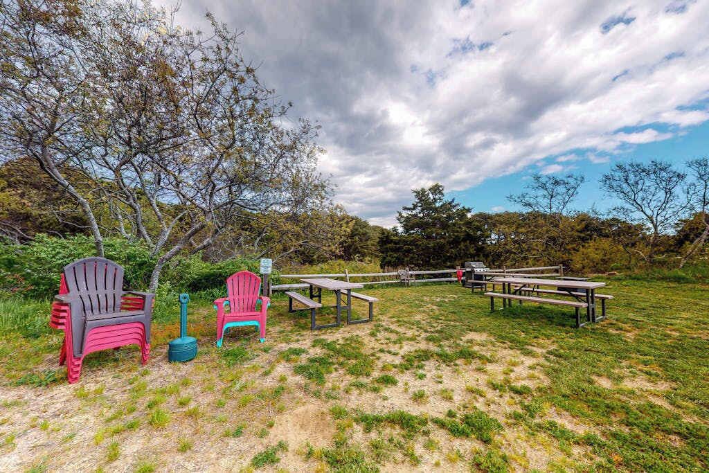 An outdoor seating area with picnic tables and chairs, surrounded by trees and grass.