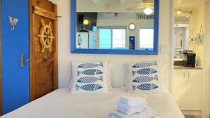 A bedroom with a bed, two pillows with fish patterns, a wooden door with a ship's wheel, and a mirror reflecting a kitchen area.