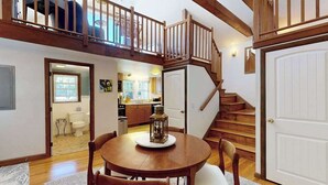 A dining area with a wooden table and chairs, a staircase with wooden railings, and a view of a kitchen and bathroom in the background. - The Lodge | Condo in Provincetown with harbor views & two decks (Provincetown)