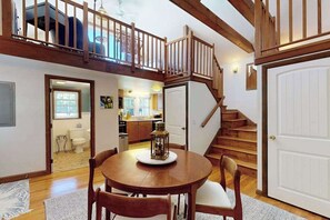 A dining area with a wooden table and chairs, a staircase with wooden railings, and a view of a kitchen and bathroom in the background. - The Lodge | Condo in Provincetown with harbor views & two decks (Provincetown)
