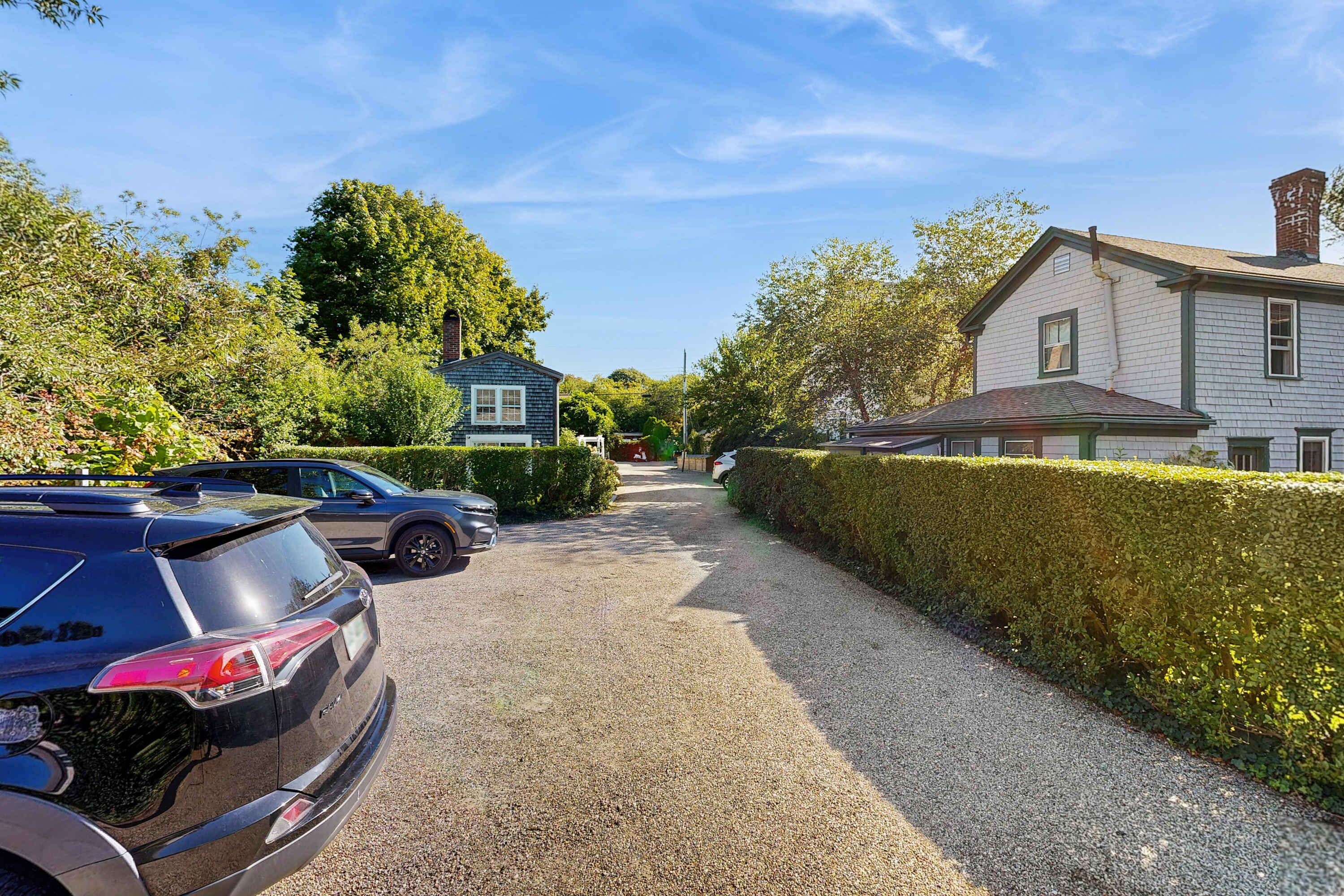 A residential street with parked cars, a gravel driveway, and houses with distinct architectural styles.