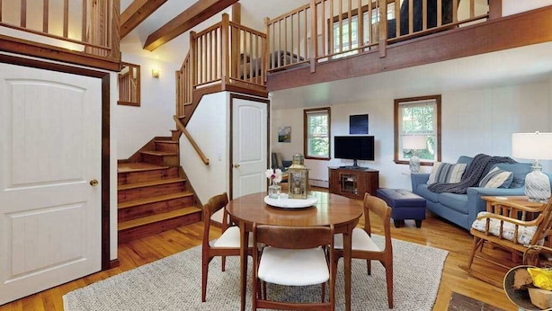 A spacious living area with a wooden staircase, a dining table with chairs, a blue sofa, and a television. - The Lodge | Condo in Provincetown with harbor views & two decks (Provincetown)
