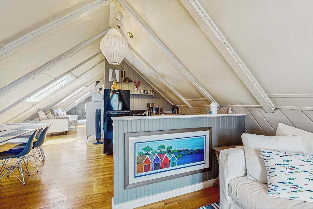 An attic bedroom with a loft bed, a dining area with a table and chairs, a kitchen area, and a large window.