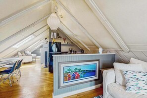 An attic bedroom with a loft bed, a dining area with a table and chairs, a kitchen area, and a large window.