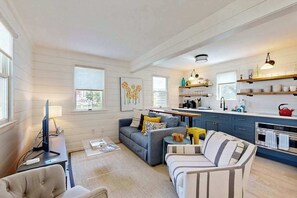 Ein modernes Wohnzimmer mit Sofa, Sessel und Couchtisch, eine Küche mit blauen Schränken und einem roten Wasserkocher sowie ein Essbereich mit Tisch und Stühlen.
