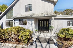 A modern two-story house with a dark front door, large windows, and a well-maintained front yard with shrubs and planters. - Spacious island home near Farm Neck Golf Course, the beach and town (Oak Bluffs)