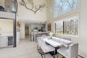 A modern kitchen with white cabinets, a dining area with a white table and chairs, and a large window with a view of trees. - Spacious island home near Farm Neck Golf Course, the beach and town (Oak Bluffs)