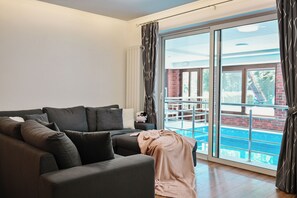 Ein modernes Wohnzimmer mit einem dunkelgrauen Sofa, einer Decke und Blick auf einen Swimmingpool durch große Glastüren.