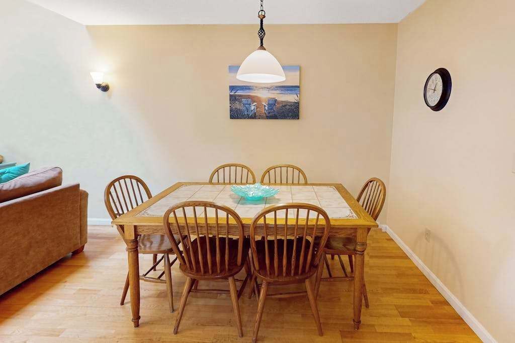 Een eetruimte met een houten tafel, zes stoelen, een blauwe schaal, een hanglamp, een schilderij en een klok aan de muur.