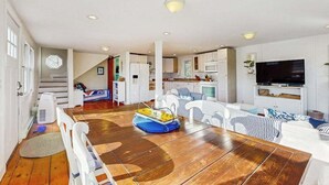 Een ruime woonkamer met een houten eettafel, witte meubels, een keuken op de achtergrond en een aan de muur bevestigde televisie.