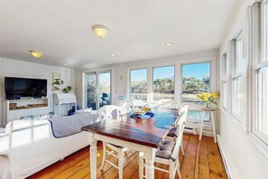 Una habitación amplia con una mesa de comedor de madera, muebles blancos, televisor y ventanas grandes que ofrecen vistas al exterior.