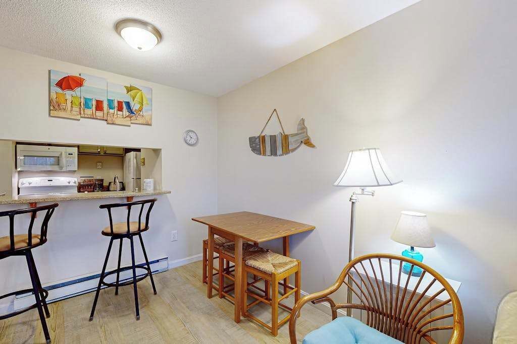 A small kitchen with a microwave, a sink, and a bar area with stools. There is a dining table with chairs and a lamp.