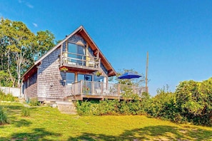 A two-story wooden cabin with a balcony, surrounded by greenery. - Water-view home with large deck, balcony, enclosed sunroom & peaceful location (Aquinnah)