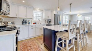 A modern kitchen with white cabinets, a central island, and a dining area with bar stools. - Beautifully renovated home near Scargo Lake with deck, W/D, AC, games, & yard (Dennis)