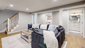A modern living room with a dark sofa, a coffee table, a staircase, and a kitchen area with white cabinets. - Beautifully renovated home near Scargo Lake with deck, W/D, AC, games, & yard (Dennis)