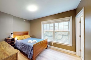A bedroom with a wooden bed, a nightstand, a desk, and a large window with a stone wall view.