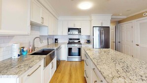 A modern kitchen with white cabinetry, a granite countertop, stainless steel appliances, and a wooden floor. - Sleek, modern home with central AC - steps to private beach & near trail (Dennis Port)