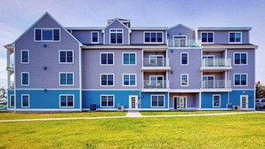A multi-story residential building with a flat roof, multiple balconies, and a well-maintained lawn in front. - Sleek, modern home with central AC - steps to private beach & near trail (Dennis Port)